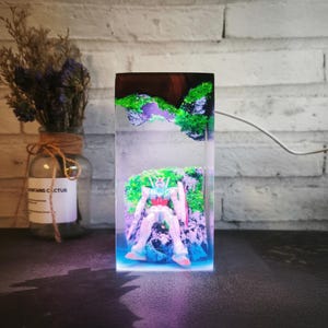May include: A rectangular resin sculpture featuring a robot figure set against a backdrop of miniature green and purple landscapes. The sculpture is illuminated with a blue and purple light, creating a vibrant display. The robot has red, white, and blue accents.