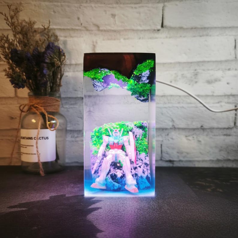 May include: A rectangular resin sculpture featuring a robot figure set against a backdrop of miniature green and purple landscapes. The sculpture is illuminated with a blue and purple light, creating a vibrant display. The robot has red, white, and blue accents.