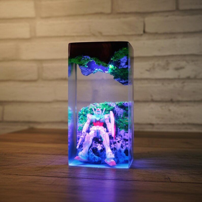 May include: A rectangular resin art piece with a robot figure in a miniature landscape. The scene includes green moss, blue lighting, and a dark top, creating a unique, illuminated display. The robot is white, red, and blue.
