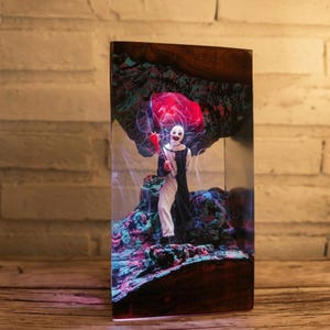 May include: A rectangular resin and wood lamp featuring a miniature clown figure in a black and white suit holding a red object. The interior displays a textured, multi-coloured landscape with swirling white smoke effects and a glowing red light.