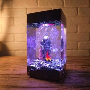 May include: A rectangular resin sculpture featuring a seated figure with a campfire scene. The sculpture is encased in clear resin with swirling purple and white accents. The base is dark, and the figure is detailed with a small fire.