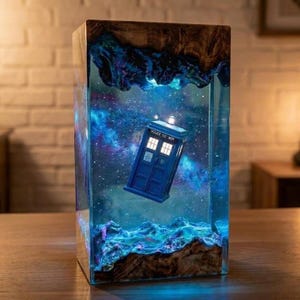 May include: A rectangular lamp featuring a blue police box suspended within a clear resin block. The resin contains a swirling nebula of blue and purple hues with white star-like specks. The top and bottom edges consist of natural wood.