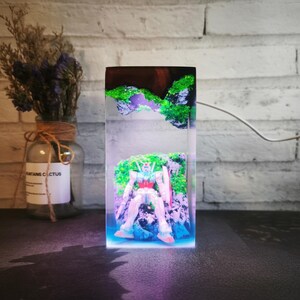 May include: A rectangular resin sculpture featuring a robot figure set against a backdrop of miniature green and purple landscapes. The sculpture is illuminated with a blue and purple light, creating a vibrant display.