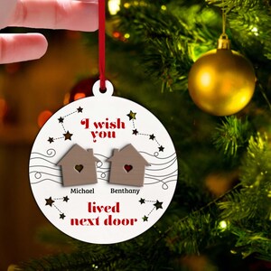 以下が含まれることがあります： 「I wish you lived next door」の文字が入った、丸い白いクリスマスオーナメント。ハートの切り抜きがある小さな木製の家が2つ、その下にあります。「Michael」と「Benthany」の名前が下にあります。赤いリボンで飾られています。