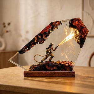 Könnte beinhalten: Eine dekorative Skulptur aus Harz und Holz mit einer Miniatur-Feuerwehrperson in voller Ausrüstung, die einen Schlauch hält. Die Figur befindet sich zwischen zwei dunklen, verkohlten Holzstücken in Harz mit einem Lichteffekt, der Feuer simuliert.