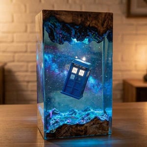 May include: A rectangular resin lamp featuring a blue police box floating in a nebula-like space scene. The lamp is encased between two pieces of natural wood with a rough edge, and the interior glows with blue and purple light.