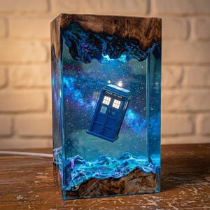 May include: A rectangular lamp featuring a blue police box model suspended within a clear resin block. The resin contains a swirling blue and purple nebula design with embedded lights. The top and bottom are framed by natural wood with live edges.