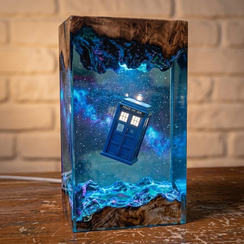May include: A rectangular lamp featuring a blue police box model suspended within a clear resin block. The resin contains a swirling blue and purple nebula design with embedded lights. The top and bottom are framed by natural wood with live edges.