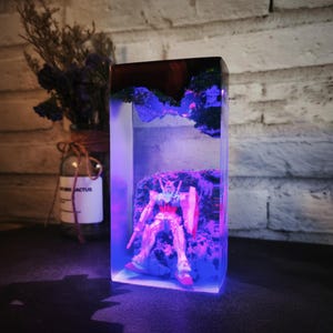 May include: A rectangular resin sculpture with a Gundam model inside. The sculpture has a dark wood top and a blue-lit interior. The Gundam is on a rocky base, with a shield and weapon. The clear resin allows the model to be seen. The sculpture is approximately 15 cm tall.
