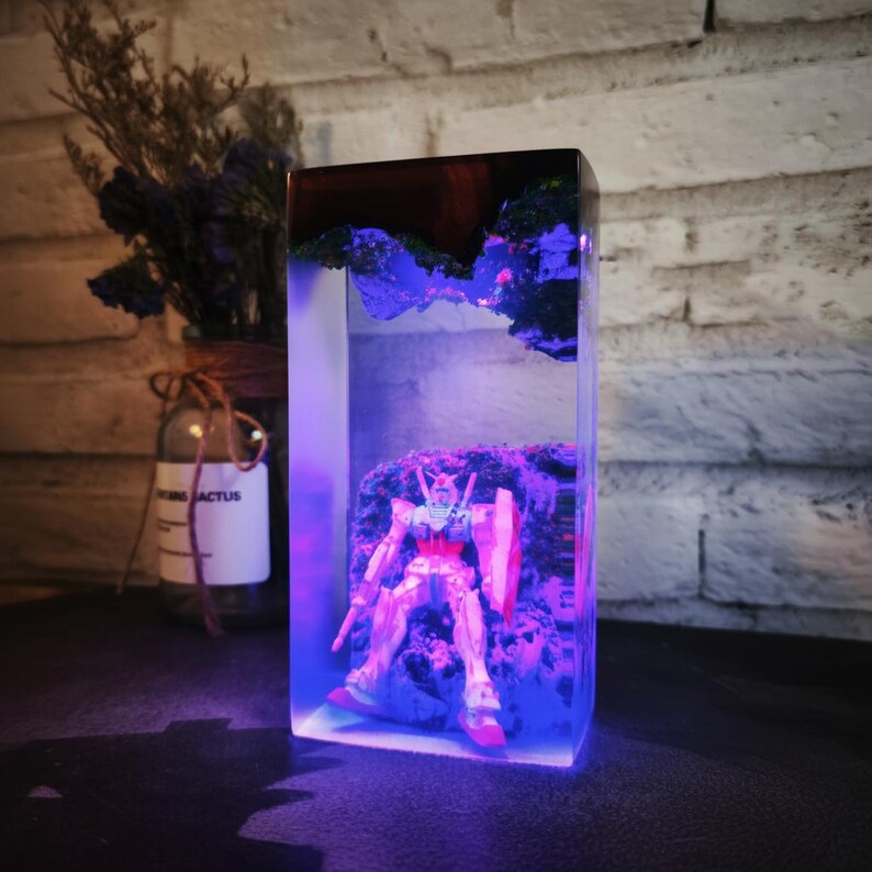 May include: A rectangular resin sculpture with a Gundam model inside. The sculpture has a dark wood top and a blue-lit interior. The Gundam is on a rocky base, with a shield and weapon. The clear resin allows the model to be seen. The sculpture is approximately 15 cm tall.