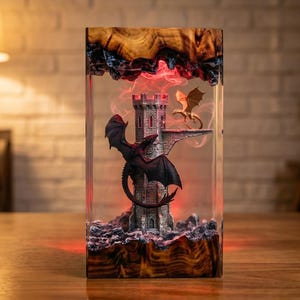 Pu&ograve; includere: Una lampada rettangolare in resina con una torre centrale color pietra, un drago scuro appollaiato sul lato e un drago dorato pi&ugrave; piccolo su una piattaforma. La scena &egrave; racchiusa in resina trasparente con venature del legno sopra e sotto.