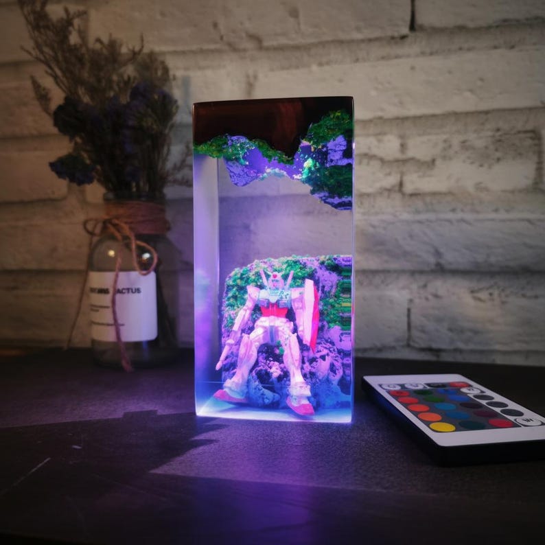 May include: A rectangular resin art piece with a robot figure set against a backdrop of simulated terrain. The resin block has a dark wood top and is illuminated with purple and green LED lights. A remote control with colour options sits nearby.