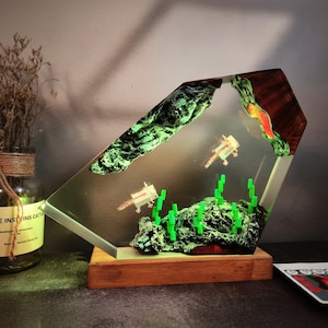 Könnte beinhalten: Ein einzigartiges Kunstwerk aus Harz mit einem klaren, abgewinkelten Design und einem Holzsockel. Es enthält zwei pixelige Axolotl, grüne Pflanzen und eine leuchtend grüne Felsformation. Die obere rechte Ecke hat eine rötlich-braune Holzmaserung.