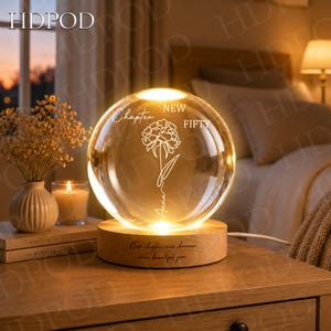 Puede incluir: Una luz nocturna esférica de cristal con un diseño floral grabado y el texto "Chapter NEW FIFTY" junto al nombre "Anna". La esfera de cristal descansa sobre una base circular de madera grabada con el texto "New chapter, new dreams, same beautiful you."