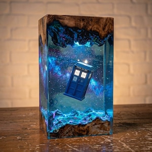 May include: A rectangular lamp featuring a blue police box suspended within a clear resin block. The resin contains a swirling blue and purple nebula effect with white star-like specks. Natural wood edges frame the top and bottom of the piece.