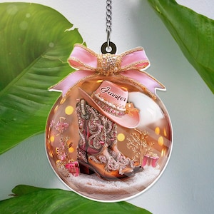 May include: A clear Christmas bauble with a pink bow and a silver chain. Inside, a pair of brown cowboy boots and a pink hat with the name "Jennifer" are set in a snowy scene. The ornament has a festive, country theme.