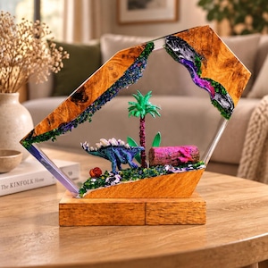 May include: A decorative display featuring a blue stegosaurus figurine, a green palm tree, and a pink log set within a geometric wooden frame with clear resin accents. The base is made of solid wood, and the scene includes small green foliage.