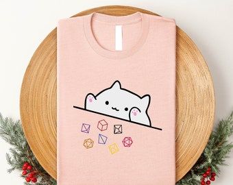Könnte beinhalten: Ein pinkfarbenes T-Shirt mit einer weißen Katze, die über den oberen Rand schaut. Die Katze hält einen Würfel in ihrer Pfote. Um die Katze herum liegen mehrere bunte Würfel verstreut.