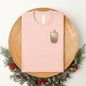 May include: A pink t-shirt with a cartoon cat in a bubble tea cup graphic on the front. The cat is brown and white with a pink straw.