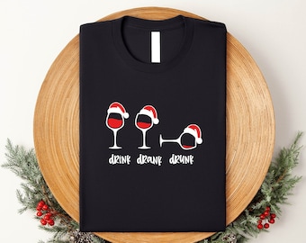 May include: Black t-shirt with a humorous graphic of three wine glasses wearing Santa hats. The glasses are labeled "drink, drank, drunk" in white text. The shirt is folded on a wooden tray with holiday greenery.