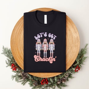 May include: Black t-shirt featuring the text "Let's Get Chackin'" and a graphic of three pastel-coloured nutcrackers. The folded shirt rests on a wooden tray adorned with greenery and red berries.