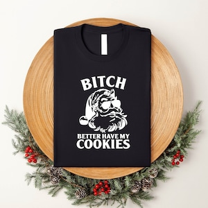 May include: Black t-shirt with a white graphic of Santa Claus wearing sunglasses and the text "BITCH BETTER HAVE MY COOKIES". The shirt is folded and placed on a wooden plate, surrounded by greenery and red berries.