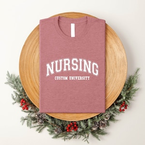 Puede incluir: Una camiseta de color rosa claro con el texto "Nursing Custom University" en blanco.