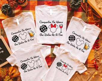 Peut inclure: Quatre t-shirts blancs avec un motif graphique noir et blanc représentant un globe terrestre, des oreilles de Mickey Mouse et une boisson. Le texte sur les t-shirts est "Conquering the World One Drink at a Time". Les boissons représentées sont un verre de vin, une bière, une brique de jus et une paille.