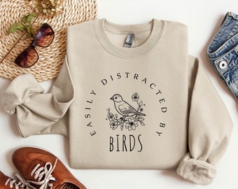 Puede incluir: Una sudadera beige con un diseño gráfico negro de un pájaro sentado en una rama con flores. El texto "Easily Distracted By Birds" está escrito en un círculo alrededor del pájaro.
