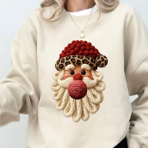 Peut inclure: Sweat-shirt beige avec un motif de Père Noël. Le visage du Père Noël a un chapeau rouge avec un bord léopard, une barbe tricotée et un nez rouge. Le motif est centré sur le devant du sweat-shirt.