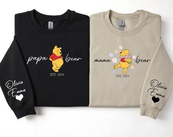May include: Two sweatshirts, one black and one tan, featuring Winnie the Pooh designs. The black sweatshirt says "papa bear" with a Pooh illustration and "EST. 2024". The tan sweatshirt says "mama bear" with a Pooh illustration and "EST. 2024".