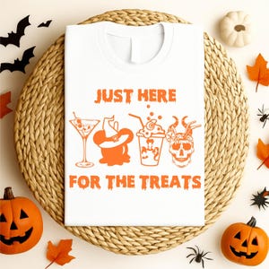 Puede incluir: Camiseta blanca con texto gráfico e ilustraciones en naranja. El texto dice "JUST HERE FOR THE TREATS". Las ilustraciones incluyen un martini, un sombrero de bruja, un batido y una calavera. Temática de Halloween.