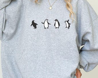 Può includere: Una felpa grigia con una grafica di quattro pinguini che camminano in fila. I pinguini sono neri e bianchi con un design semplice.