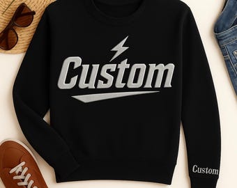 May include: Black sweatshirt with the word "Custom" in silver, featuring a lightning bolt graphic. The word "Custom" is also embroidered on the sleeve. The sweatshirt is displayed with sunglasses, a straw hat, and brown sneakers.
