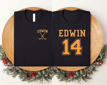 May include: Two black t-shirts with the name "EDWIN" printed in orange. One shirt has the number "14" printed in orange. The shirts are on a wooden surface.