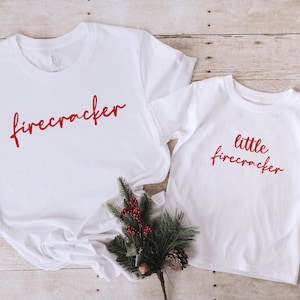 May include: Two white t-shirts with red text. The larger shirt says "firecracker" and the smaller shirt says "little firecracker".