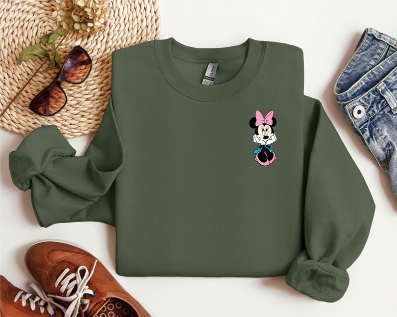Peut inclure: Un sweat-shirt vert fonc&eacute; avec une petite image brod&eacute;e de Minnie Mouse portant un n&oelig;ud rose.