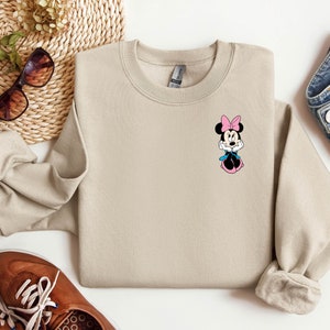 Peut inclure: Un sweat-shirt beige avec un petit graphique de Minnie Mouse sur le devant. Minnie Mouse porte un n&oelig;ud rose et une robe bleue.