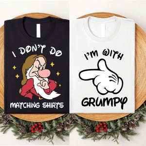 Könnte beinhalten: Zwei schwarze und weiße T-Shirts mit passendem Design. Das schwarze Shirt zeigt ein Cartoon-Bild eines Zwergs mit Bart und dem Text "Ich mag keine passenden Shirts". Das weiße Shirt zeigt eine weiße Cartoon-Hand, die nach rechts zeigt, mit dem Text "Ich bin mit Grumpy".