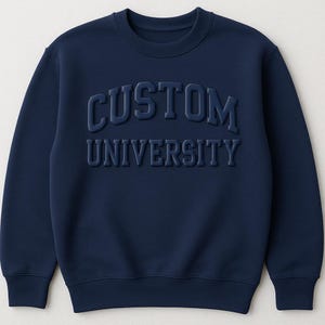 May include: Navy blue sweatshirt with the words "CUSTOM UNIVERSITY" in raised, dark blue lettering across the chest. The crew neck sweatshirt is a solid color and has a classic, casual style.