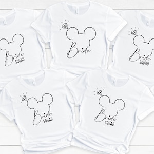 Può includere: Cinque magliette bianche con un design a testa di Topolino nero e il testo "Bride Squad" stampato su ogni maglietta.