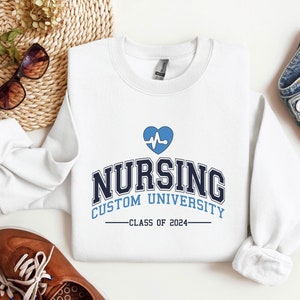 Puede incluir: Sudadera blanca con un gráfico de corazón azul y el texto "Nursing Custom University - Class of 2024".