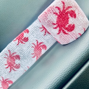May include: A white and gray fabric wrap featuring a repeating pattern of red crabs. The wrap is folded over itself, showcasing the crab design. The fabric has a textured appearance.