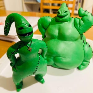 May include: Two green, stitched, cartoon figures from The Nightmare Before Christmas. The larger figure is standing with its arms raised and has a wide smile. The smaller figure is standing with its arms crossed and has a cross necklace.