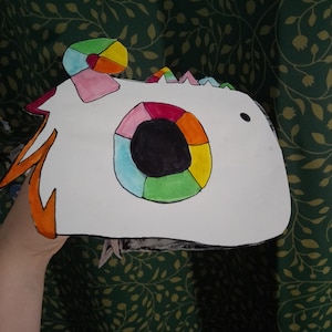 Puede incluir: Una máscara de papel blanca con un diseño de ojo colorido. El ojo está hecho de círculos en tonos de azul, verde, amarillo, naranja y rojo. La máscara tiene un pequeño remolino rosa y blanco en la parte superior.