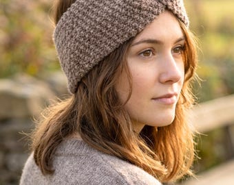 May include: A brown knitted headband with a twisted knot detail. The headband is made of a textured yarn and is worn by a person with brown hair and a matching sweater. The background is blurred, suggesting an outdoor setting.