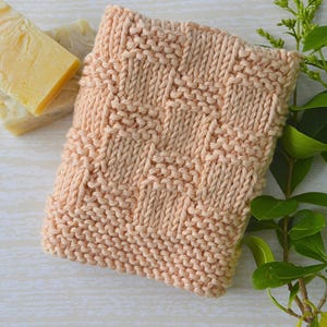 May include: A peach-colored knitted washcloth featuring a textured basketweave pattern. The square textile is positioned next to a block of pale yellow soap and a green leafy branch, showcasing the intricate knit stitches and soft yarn texture.