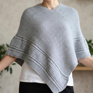 May include: A gray knitted poncho with a V-neck and flared sleeves. The poncho features textured horizontal bands and a simple, yet elegant design. The garment is made of a soft, warm material, perfect for cooler weather.