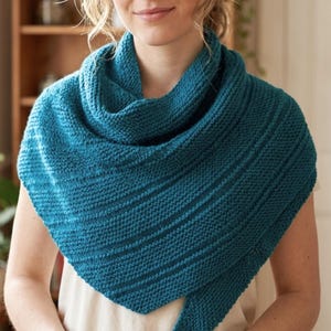 May include: A teal knitted shawl with a textured, ribbed pattern. The shawl is draped around the shoulders, forming a triangular shape. The knitted fabric has a soft appearance, suggesting warmth and comfort. The shawl is a versatile accessory.