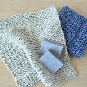 Op de afbeelding: Een lichtbeige gebreid washandje en een kleiner blauw gebreid washandje liggen naast twee rechthoekige lichtpaarse zeepblokken met zichtbare spikkels van plantaardig materiaal in het oppervlak van de zeepstukken verwerkt.
